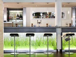A modern bar area with a counter, shelves of liquor bottles, and three bar stools. - ibis Berlin Dreilinden (Kleinmachnow)