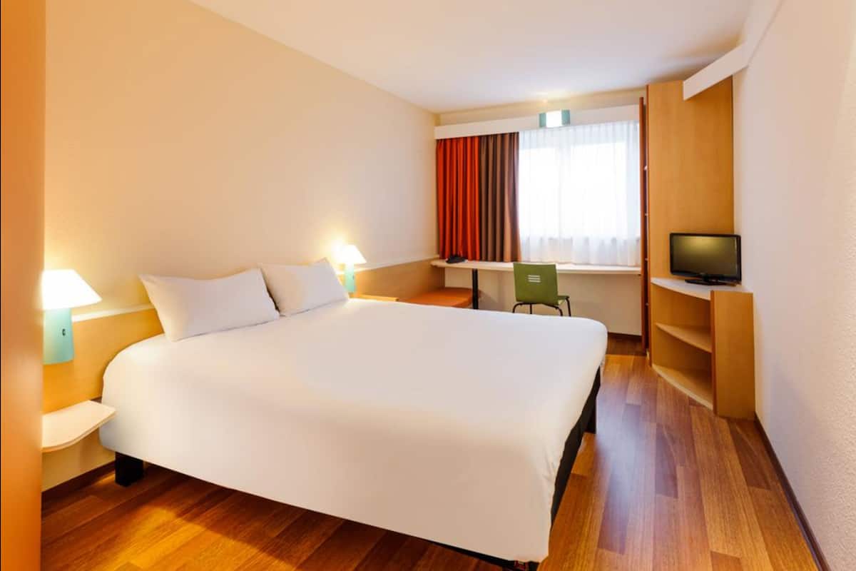A hotel room with a large bed, a desk, a chair, a television, and a window with curtains.