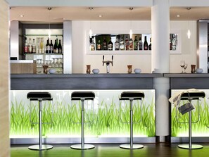 A modern bar area with a counter, shelves of liquor bottles, and three bar stools. - ibis Berlin Dreilinden (Kleinmachnow)