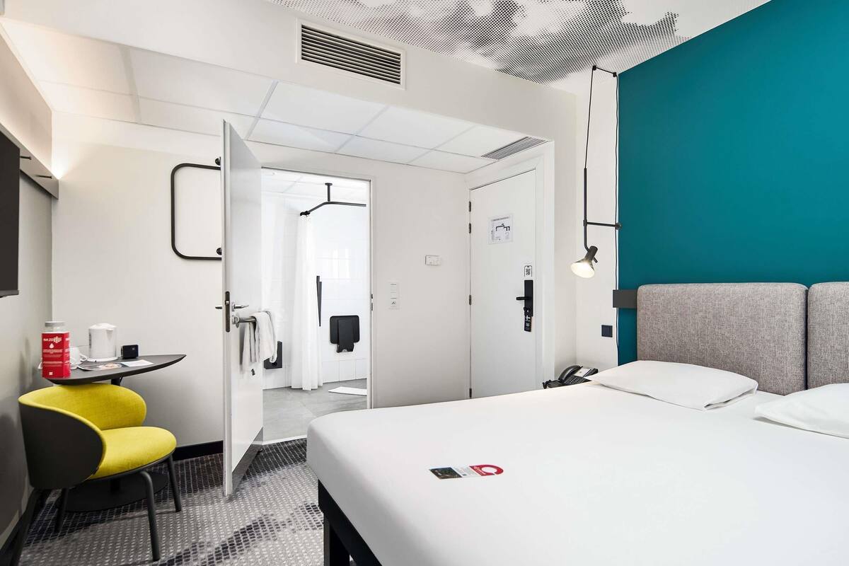 A hotel room with a bed, a desk with a chair, a bathroom visible through an open door, and a teal accent wall.