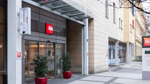 Entrada de hotel con puerta de vidrio, señalización roja y plantas en macetas.
