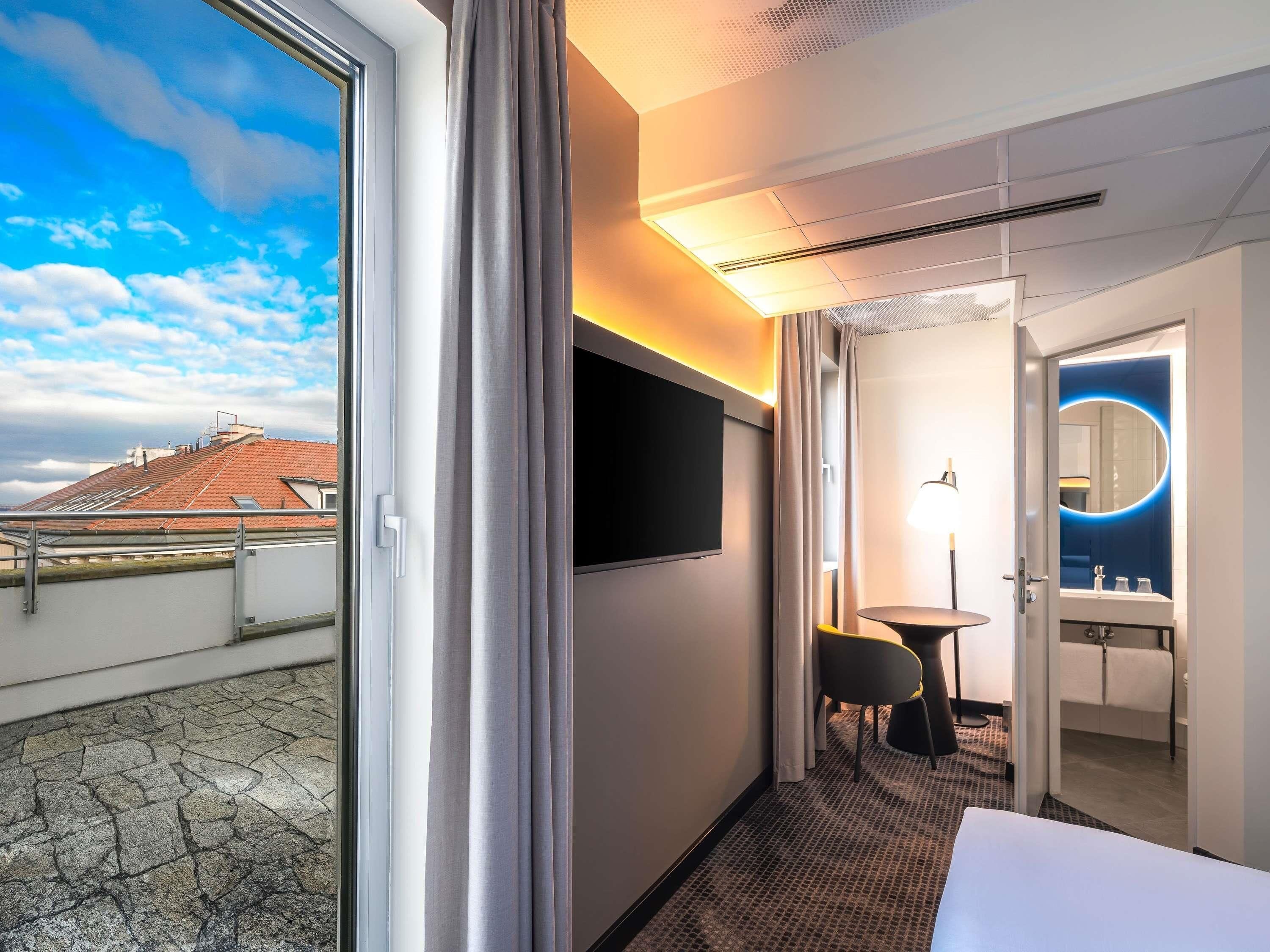 Quarto de hotel com uma grande janela, televisão de tela plana, espelho redondo, mesa pequena e uma cama.