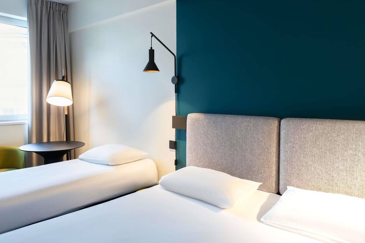 A hotel room with two beds, a nightstand, a lamp, and a view of the outdoors.