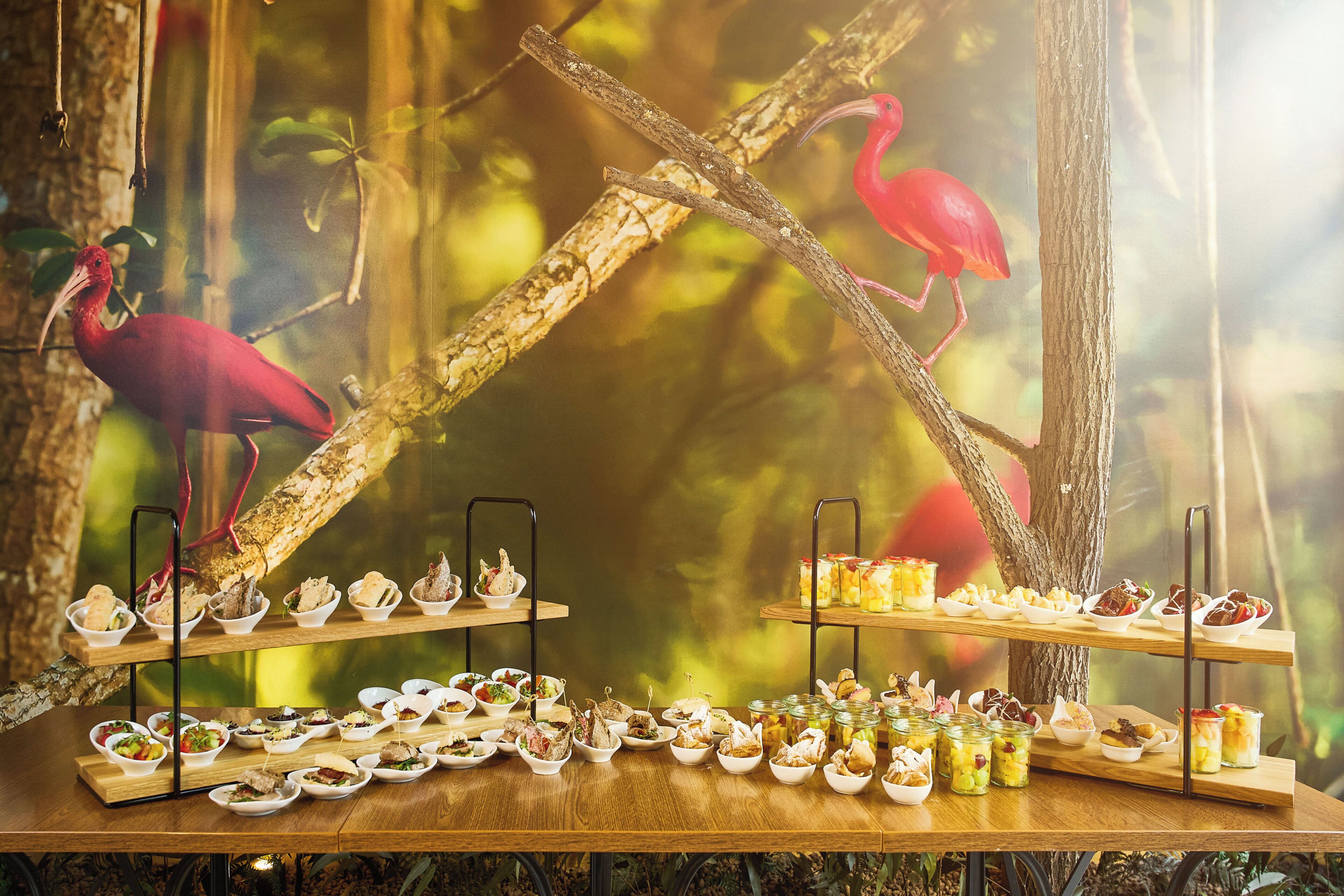 A buffet table with various dishes and a backdrop of a tree branch with a flamingo.