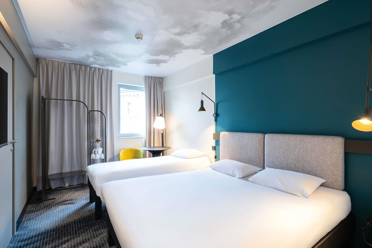 A hotel room with two beds, a window with curtains, a desk with a lamp, and a wall-mounted light fixture.