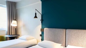 Habitación de hotel con dos camas, una mesita de noche, una lámpara y vistas al exterior.