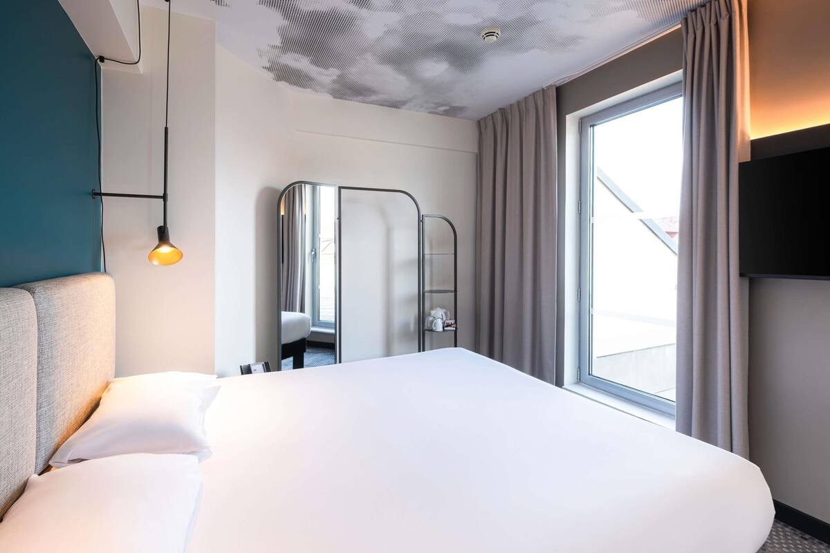 A modern hotel room with a large bed, a bedside lamp, a mirror, and a window with curtains.