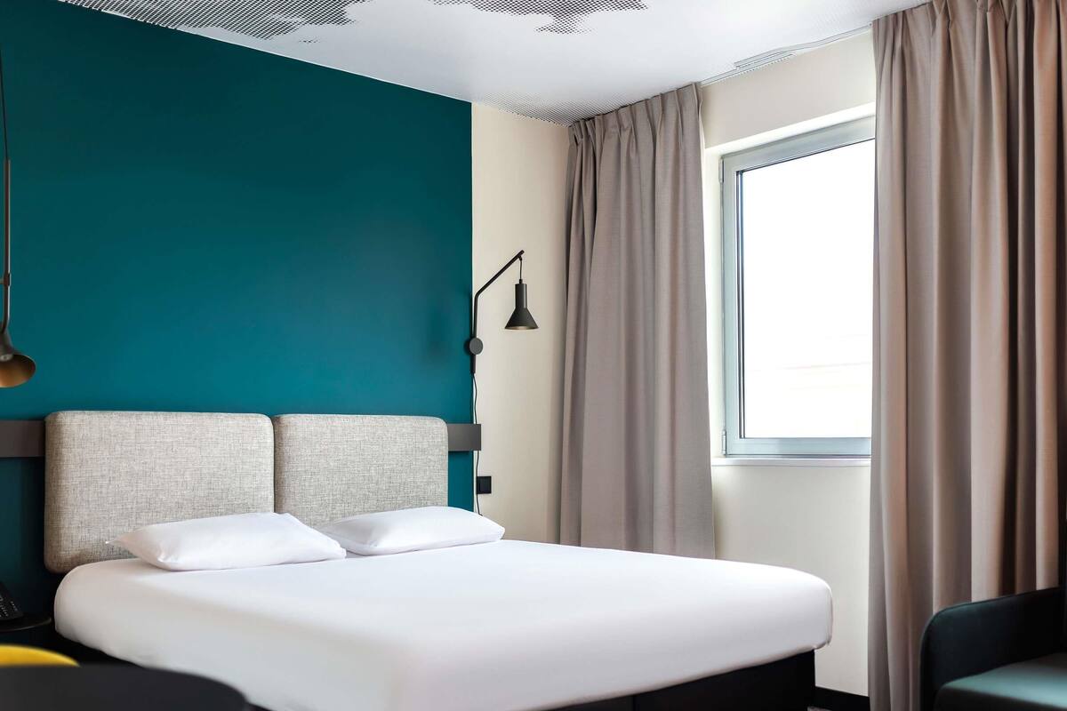 A hotel room with a bed, a desk, a lamp, and a window with curtains.