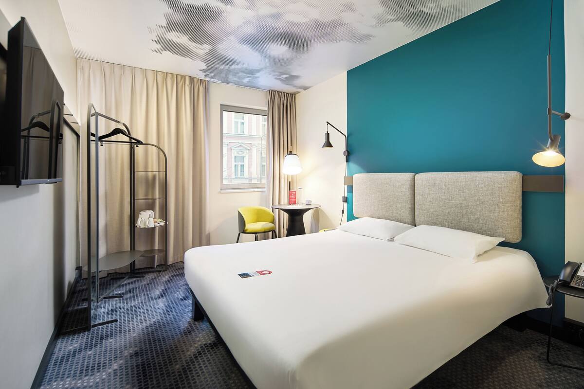 A hotel room with a large bed, a desk with a lamp, a chair, a coat rack, and a view of the outside through a window.