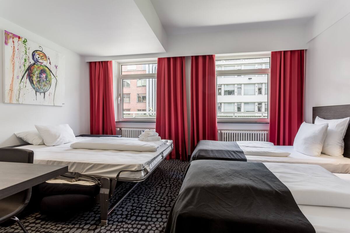 Camera d'albergo con due letti, una scrivania, una sedia e una finestra con tende rosse.