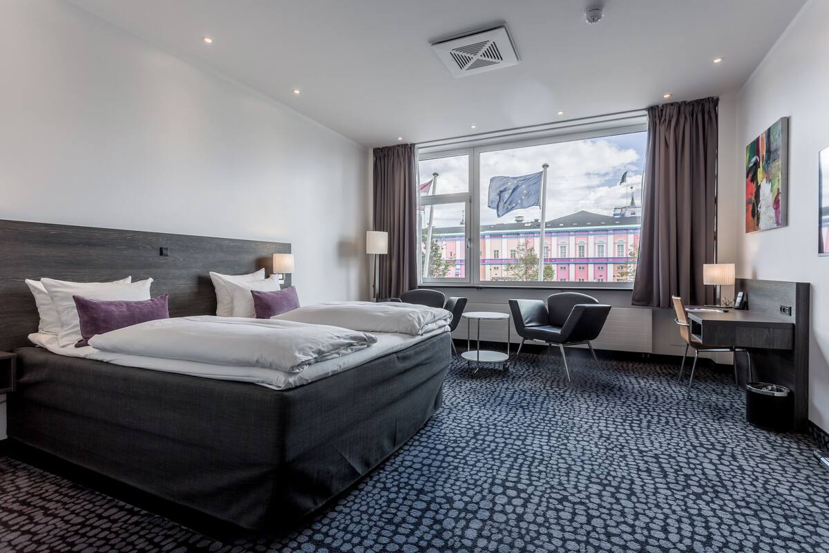 Una camera d'albergo con un letto grande, due comodini, una scrivania e una sedia. Si vede una finestra con vista su un edificio e una bandiera.