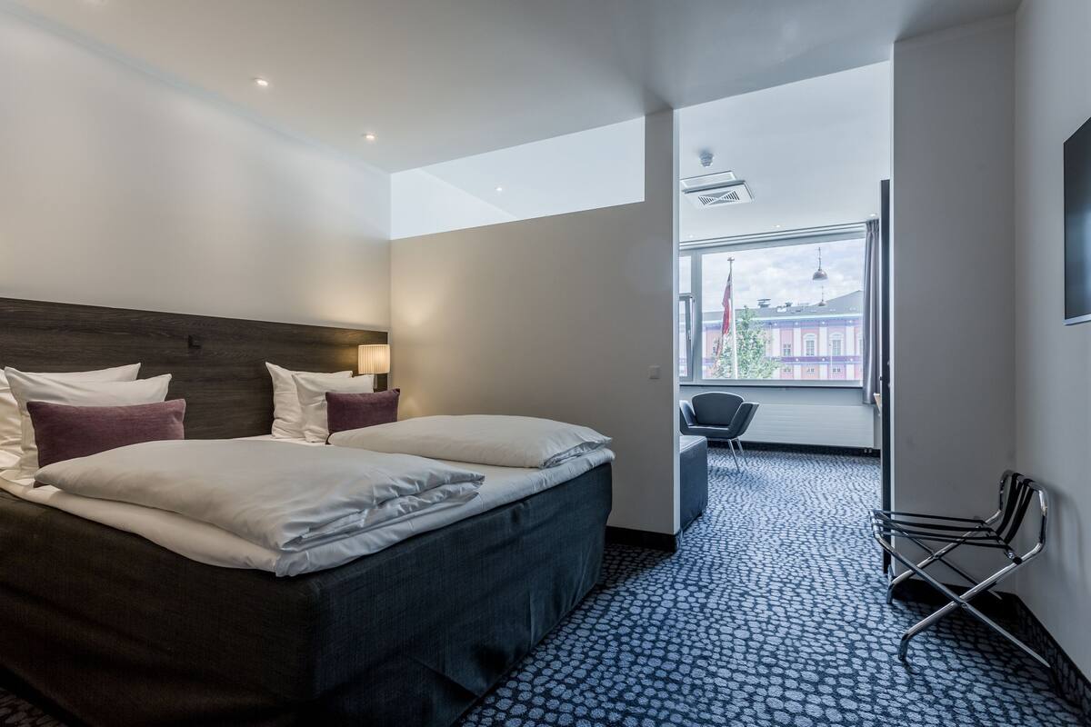 Una camera d'albergo moderna con un grande letto, una scrivania e una sedia. Si vede una finestra con vista sugli edifici.