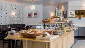 A bakery counter with bread, pastries, and fruits. There are wooden tables and chairs, and a wall with a striped pattern. - ProfilHotels Mercur (Copenhagen)