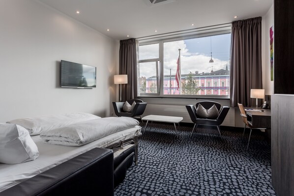 Habitación de hotel con cama, dos sillas, una mesita, un escritorio con lámpara y un ventanal con cortinas.