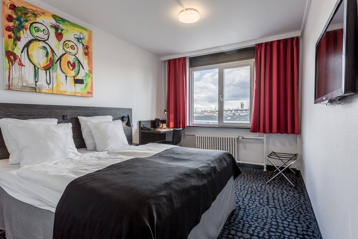 Una camera d'albergo con un letto, una scrivania, una sedia, una finestra con tende rosse, un termosifone e un quadro appeso al muro.