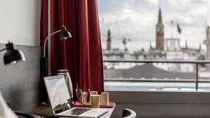 A laptop on a desk with a view of a cityscape, including a clock tower, through a window with red curtains. - ProfilHotels Mercur (Copenhagen)