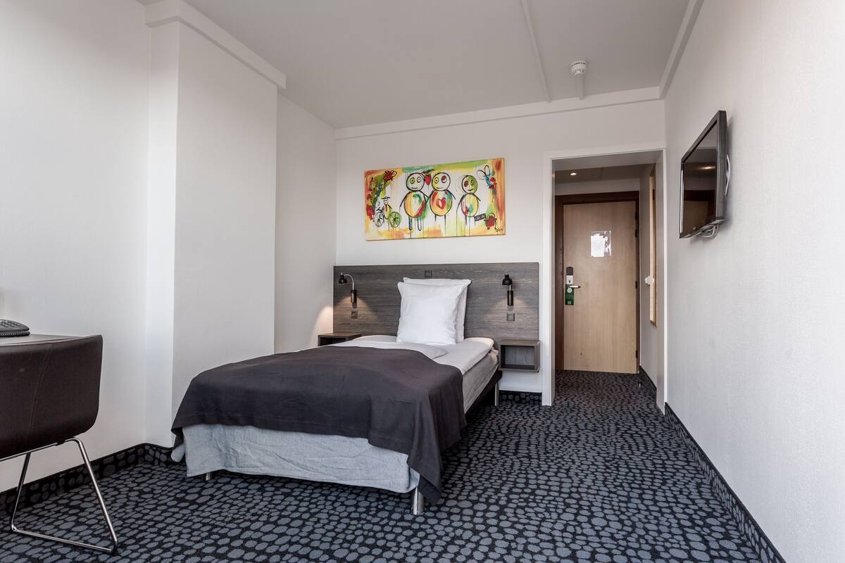 Una camera d'albergo con un letto, una scrivania, una sedia, un quadro appeso al muro e una porta che dà su un'altra stanza.