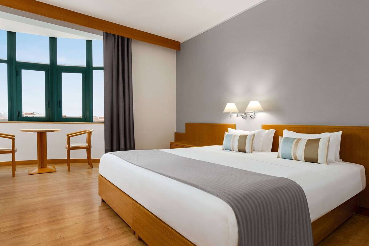Habitación de hotel con una cama grande, una mesita con dos sillas, vistas a través de amplios ventanales y una lámpara fijada a la pared.