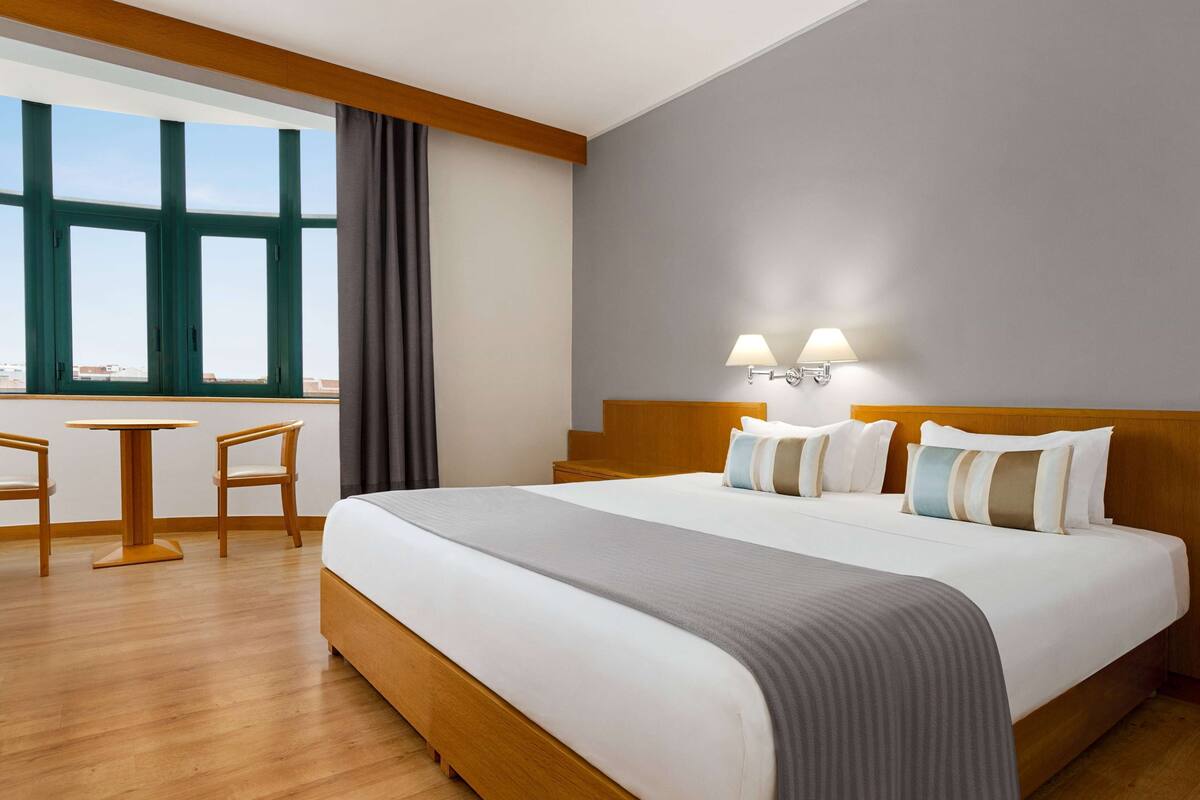 Habitación de hotel con una cama grande, dos lámparas de noche, vistas a través de ventanales del suelo al techo, una mesita y dos sillas.