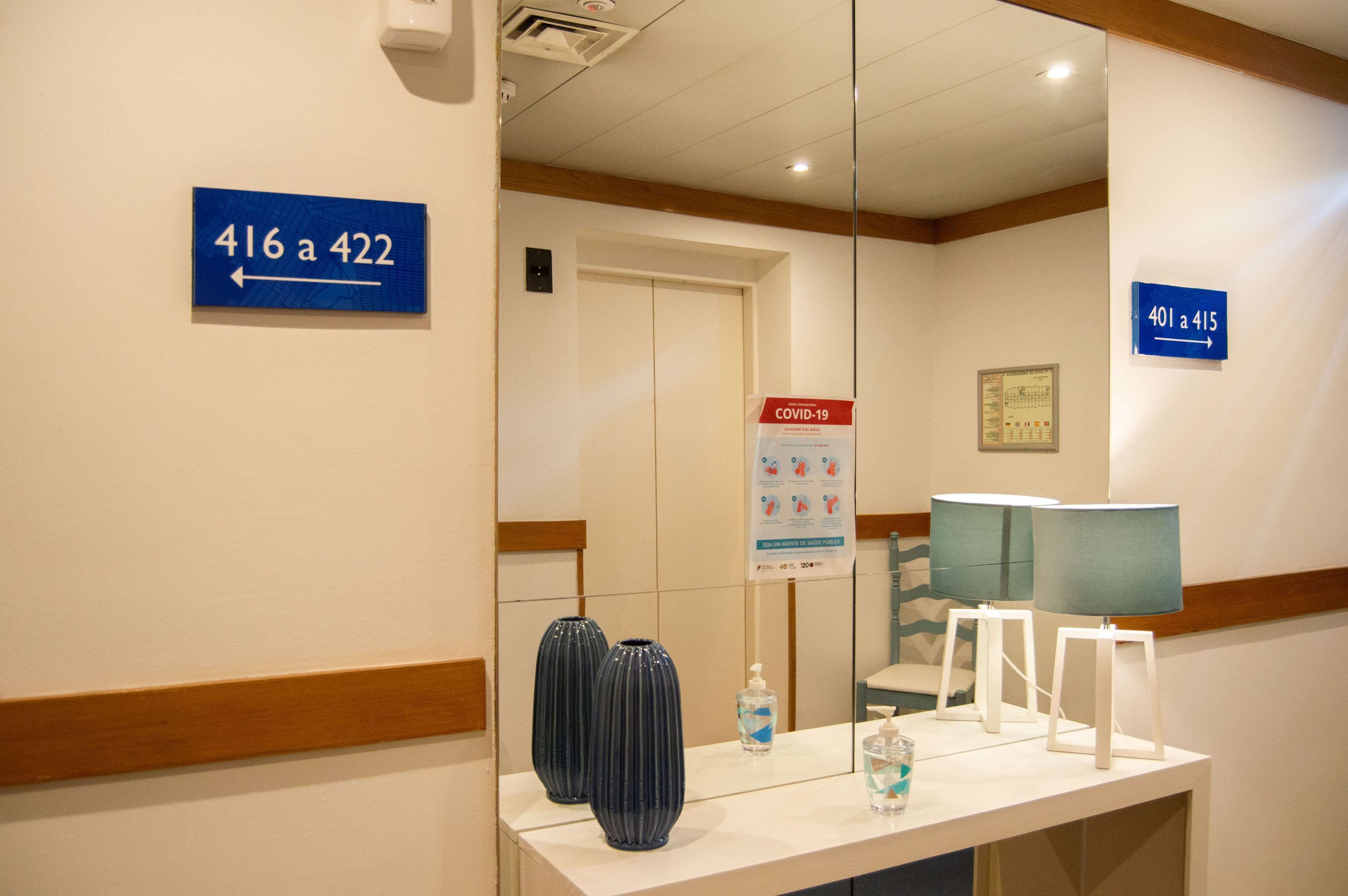 Corredor de hotel com placas de sinalização, cartaz informativo sobre COVID-19 e uma mesa de recepção com vasos e luminárias.
