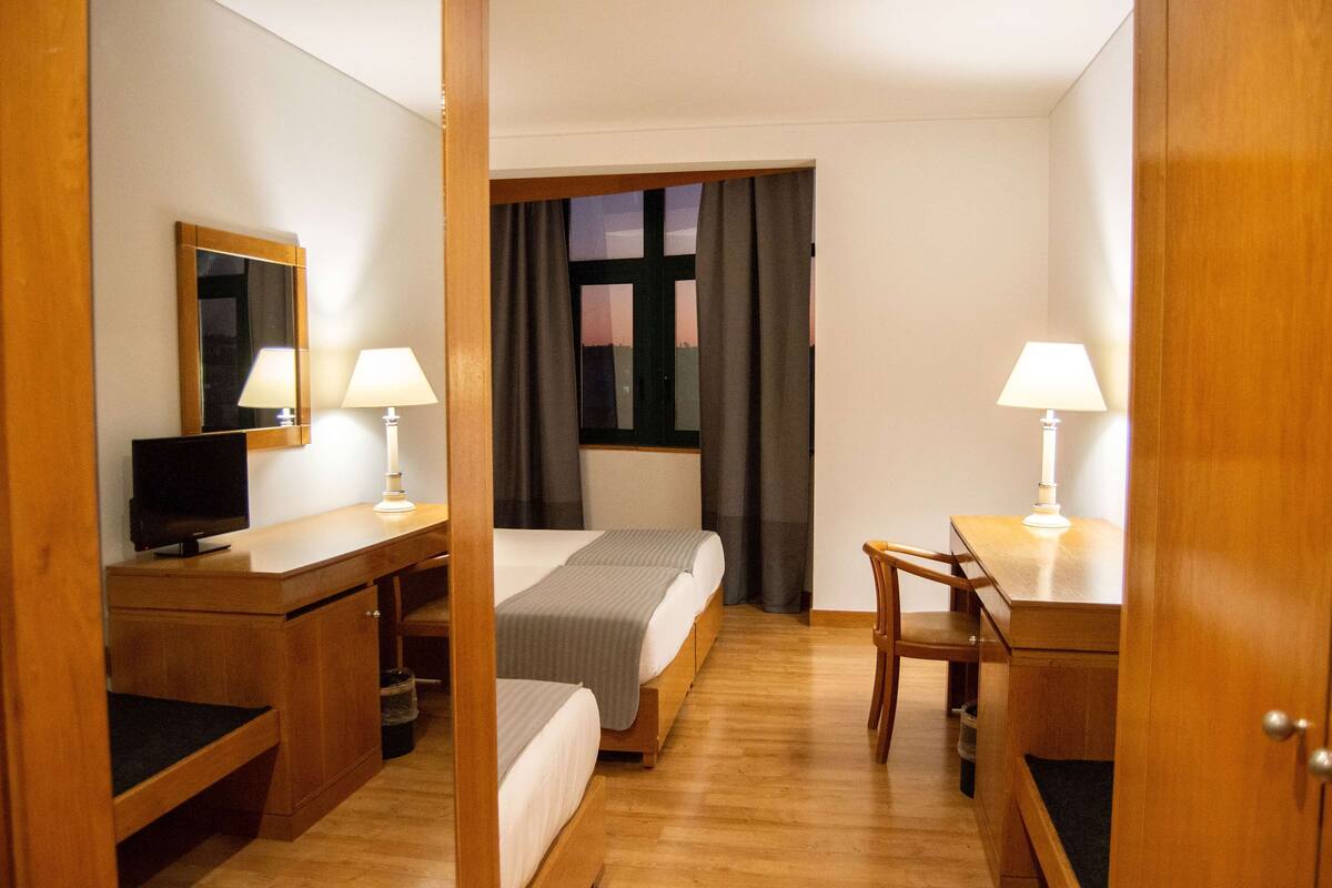 Una habitación de hotel con una cama, un escritorio con computadora, una silla y una ventana con cortinas.