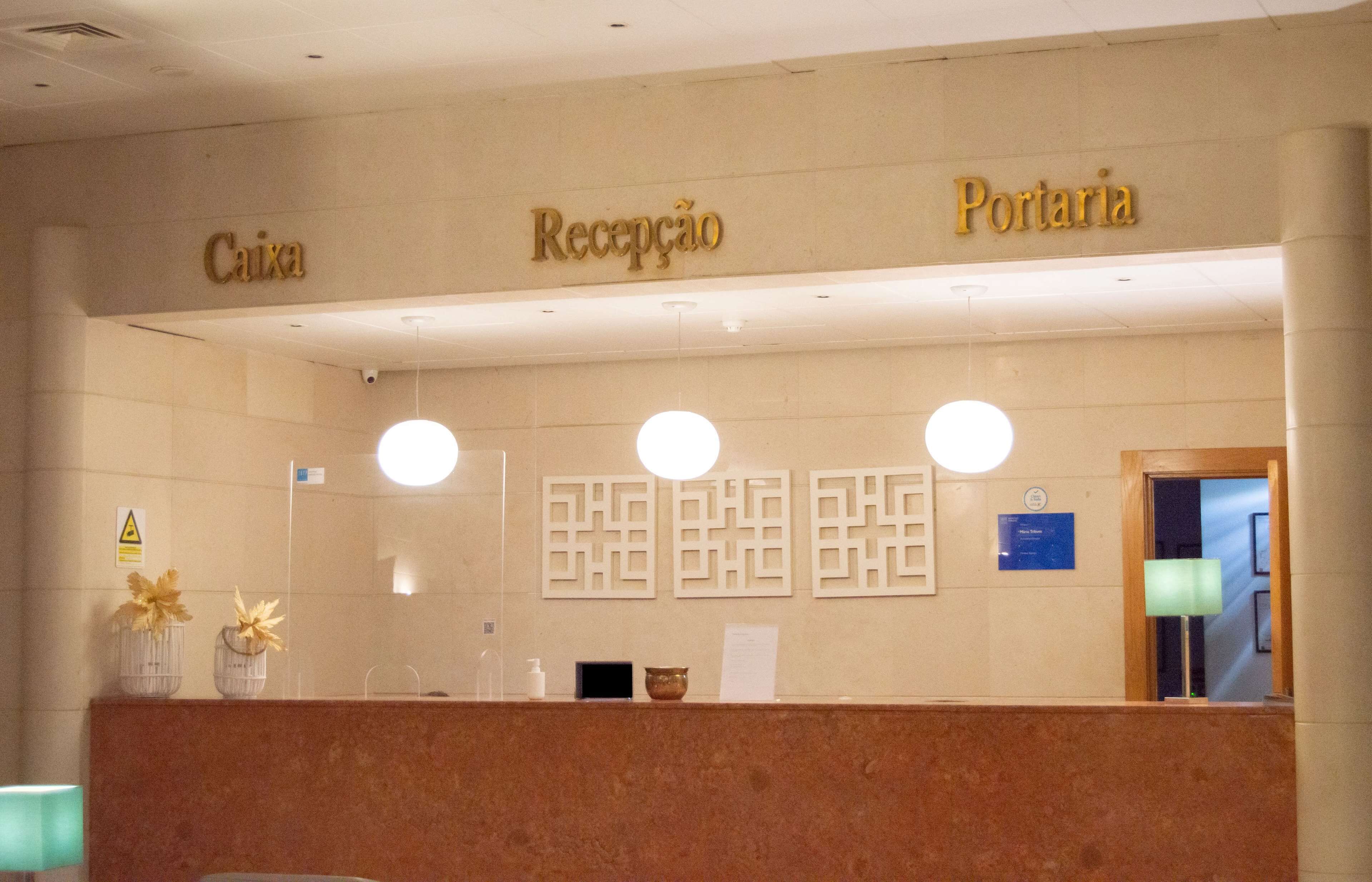A reception desk with 'Caixa' and 'Portaria' signs, a mirror, and a wall-mounted light fixture.