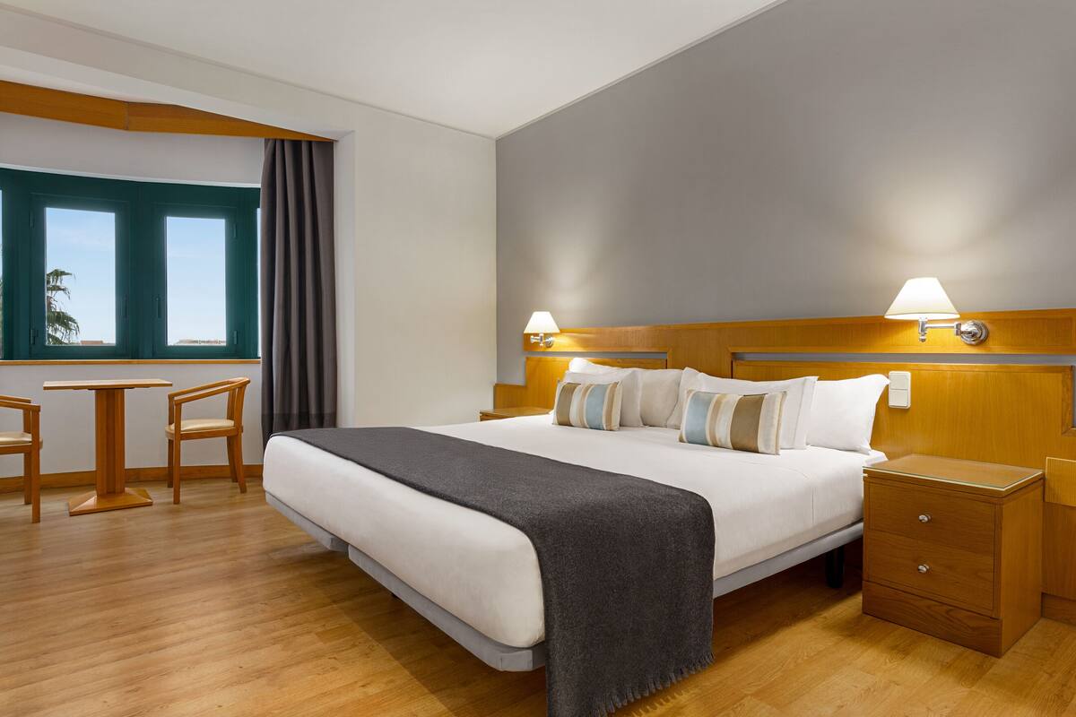 Una habitación de hotel con cama, mesitas de noche, un escritorio con sillas y una ventana con cortinas.