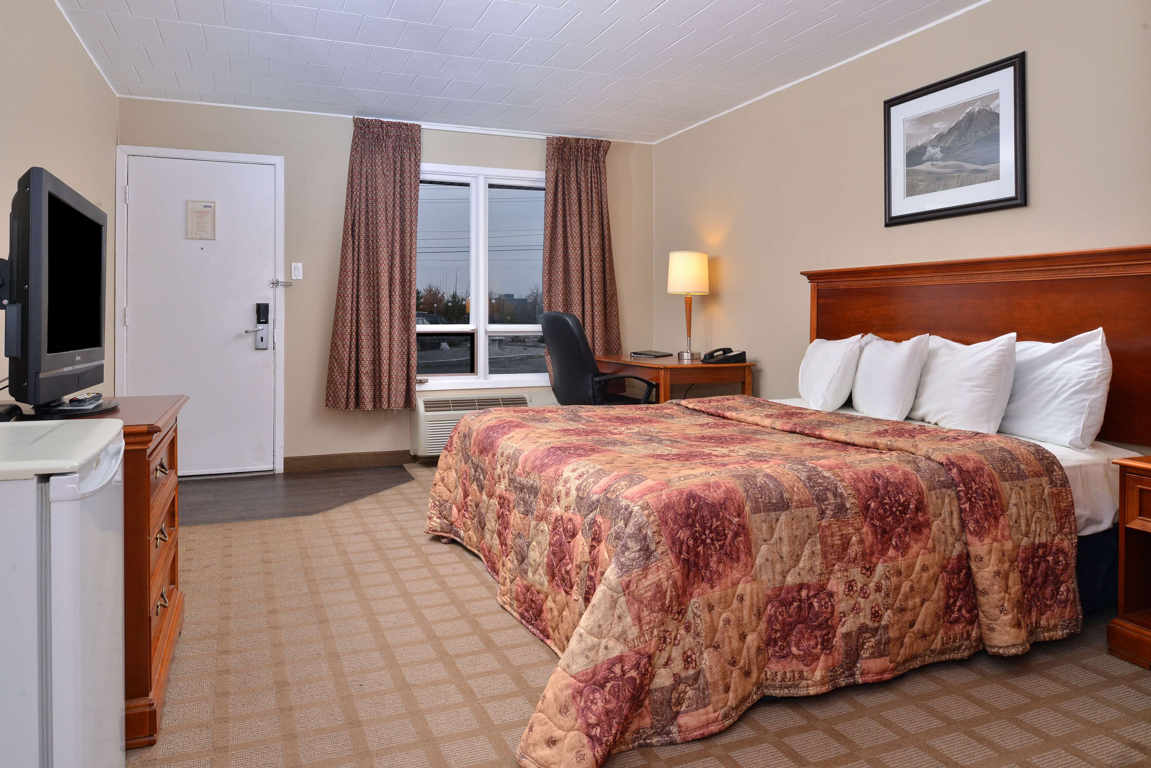 A hotel room with a large bed, a desk with a lamp, a chair, a television, and a window with curtains.