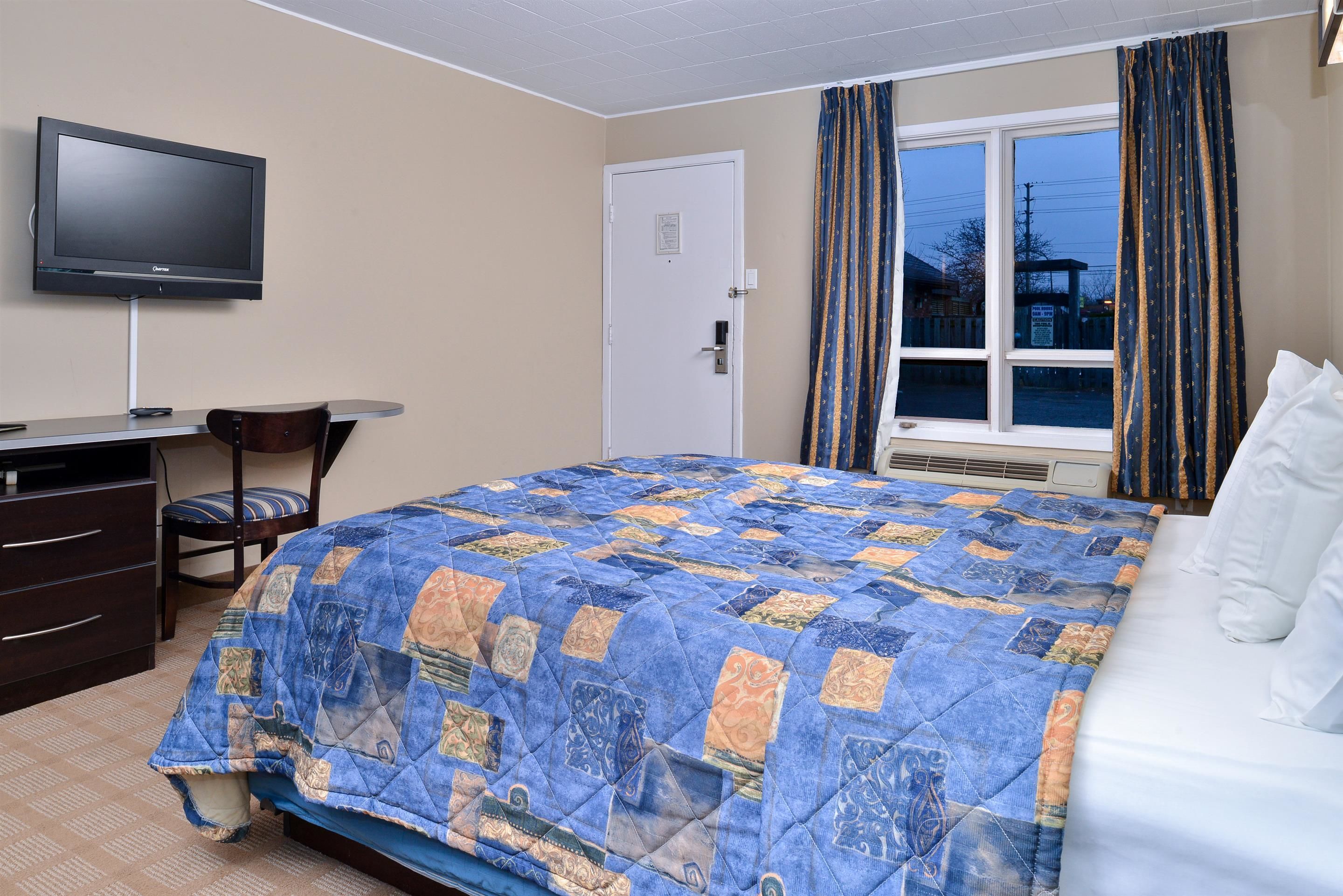 A hotel room with a bed, a desk with a chair, a television, and a window with curtains.