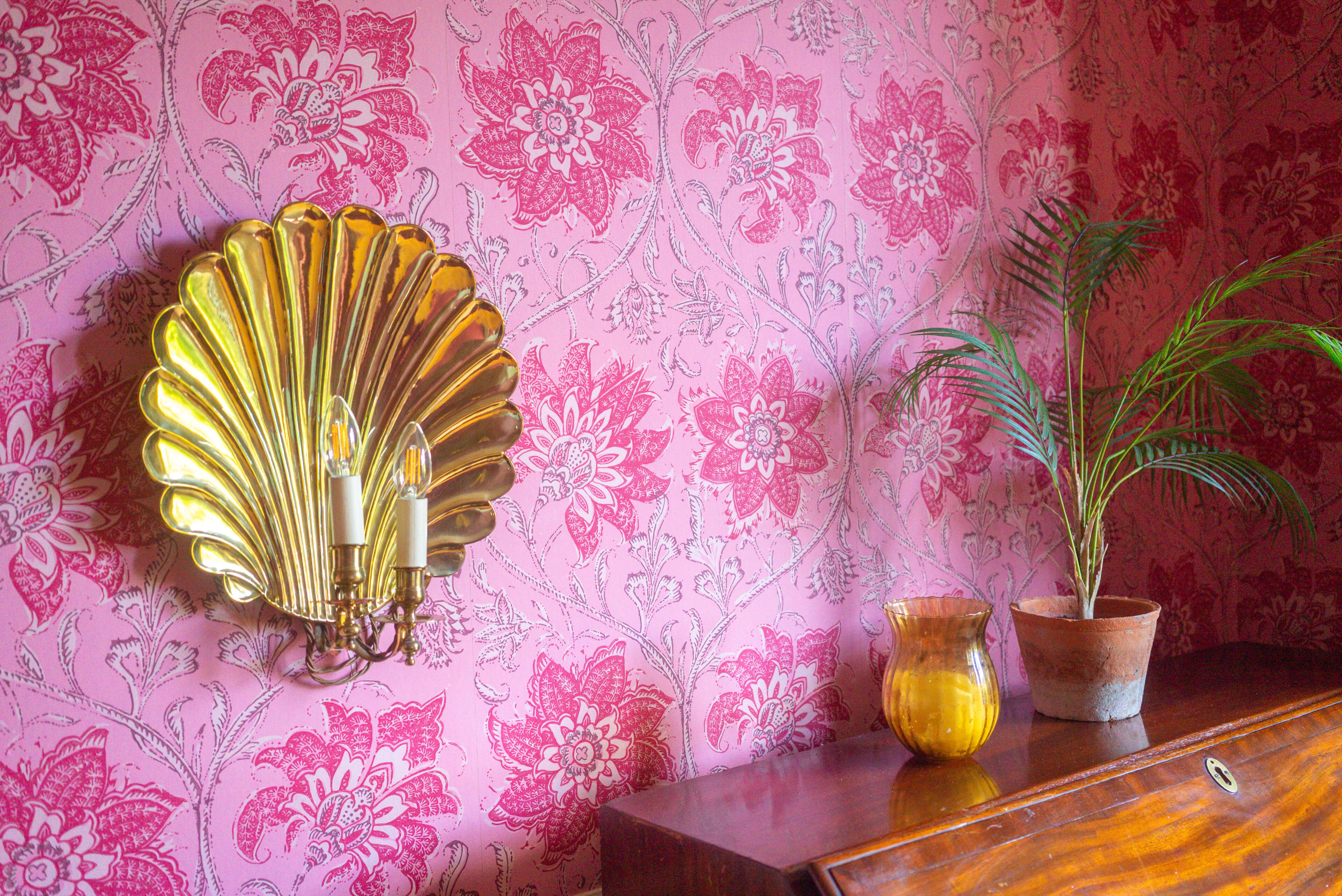 Una pared con estampado de papel tapiz floral, un aplique decorativo dorado, una planta en maceta y dos jarrones de cristal sobre un mueble de madera.