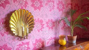 Una pared con estampado de papel tapiz floral, un aplique decorativo dorado, una planta en maceta y dos jarrones de cristal sobre un mueble de madera.