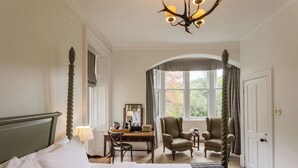 Habitación de hotel con cama, escritorio, dos sillones, una araña y un gran ventanal con cortinas.