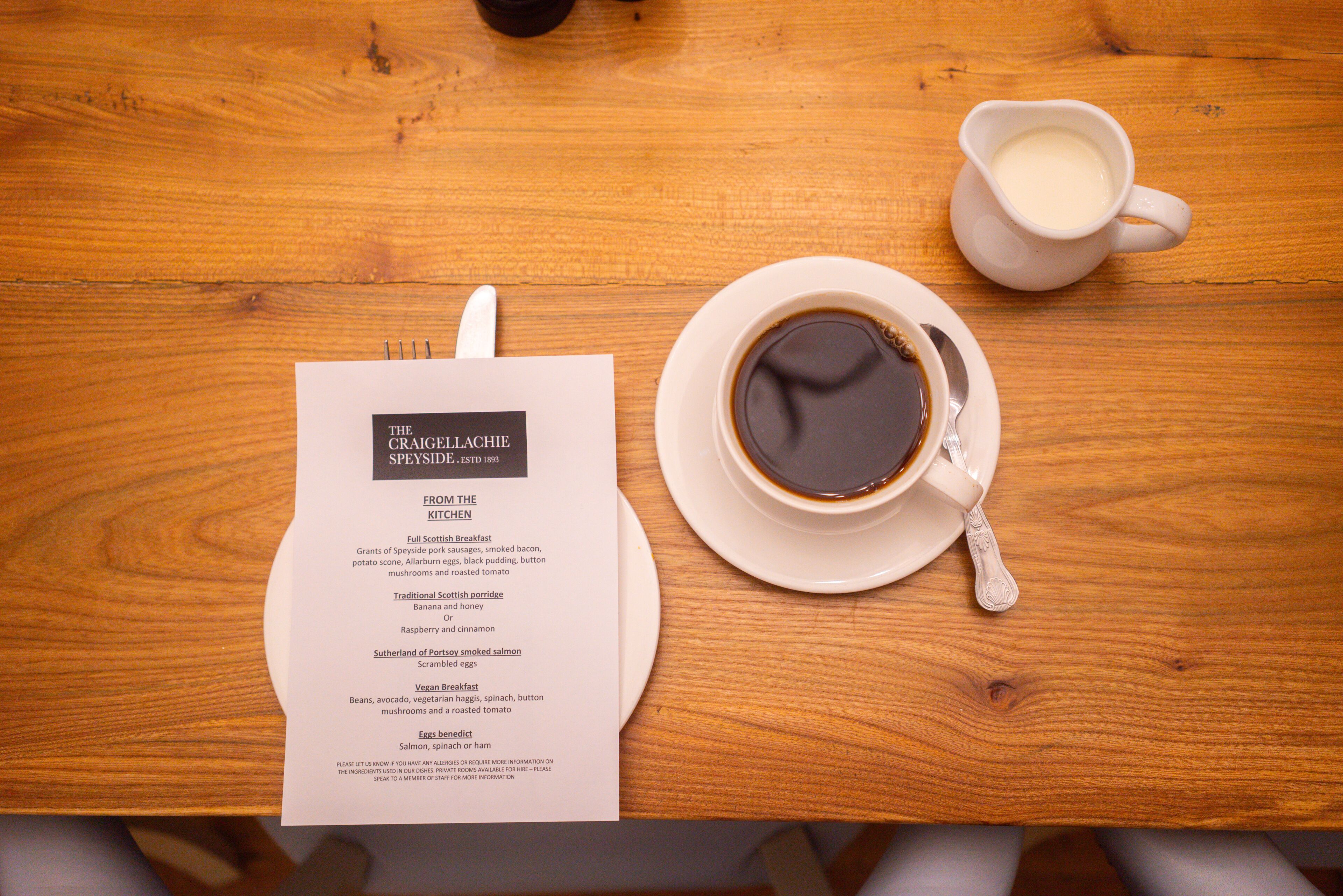 A wooden table with a menu, a cup of coffee, and a jug of milk.