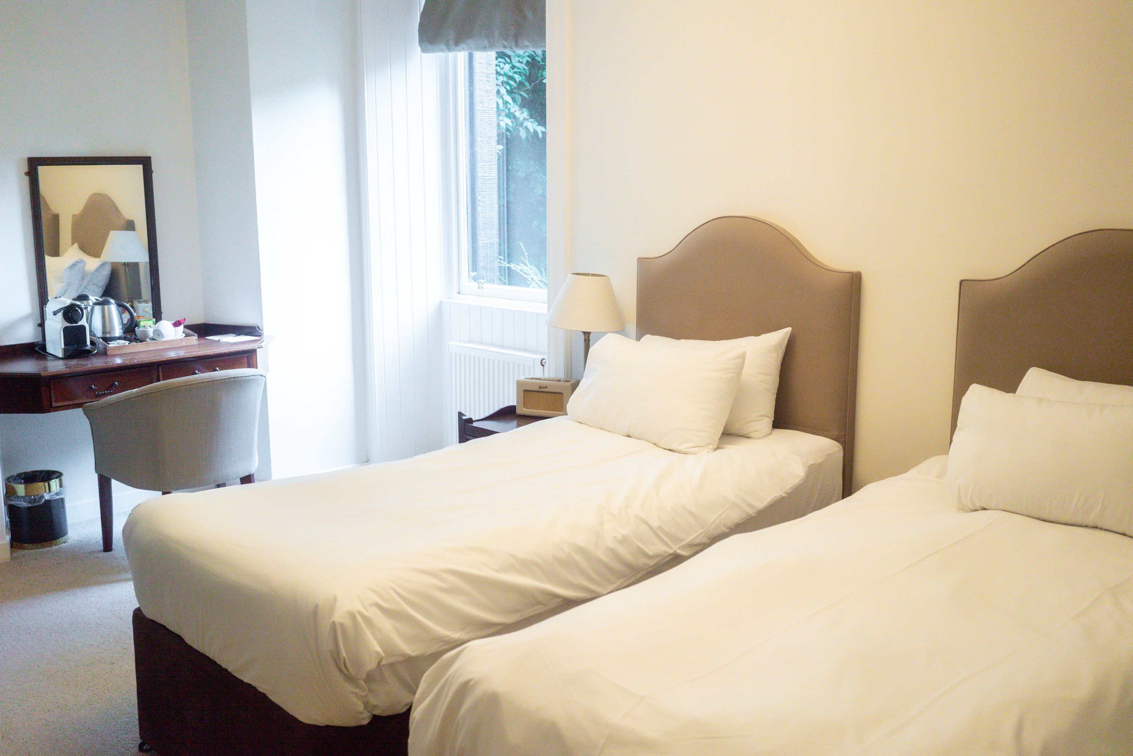 Habitación de hotel con dos camas, un escritorio con lámpara y ventana con vista a árboles.