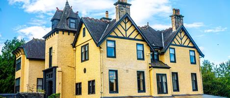 Un edificio grande y amarillo con múltiples chimeneas y marcos de ventanas negros, que cuenta con una escalera destacada y un área exterior cubierta.