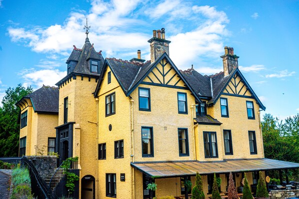 Un edificio grande y amarillo con múltiples chimeneas y marcos de ventanas negros, que cuenta con una escalera destacada y un área exterior cubierta.