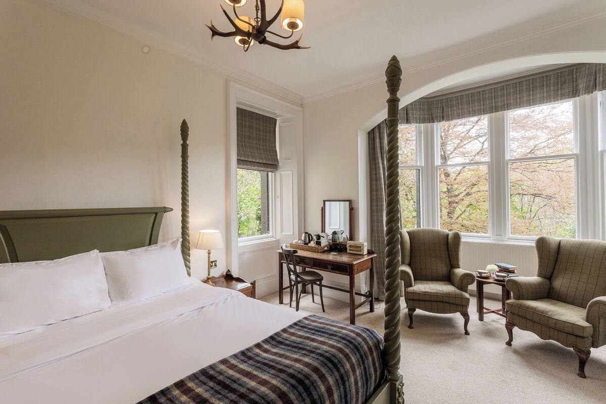 Ein Bett mit Baldachin, ein grüner Quetschkopf, ein Schreibtisch mit Stuhl, ein kleiner Tisch mit Büchern und ein großes Fenster mit Vorhängen.