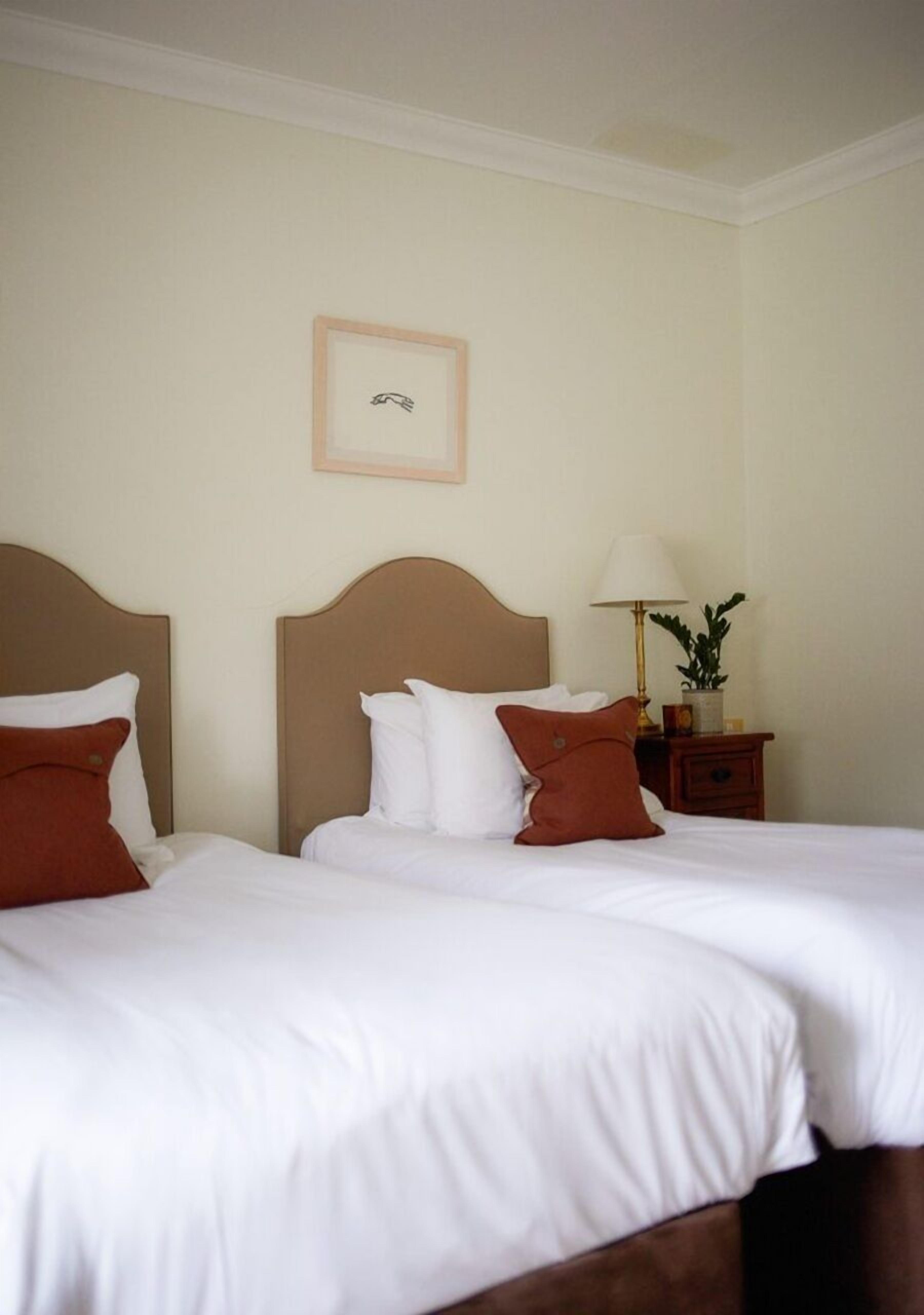 A hotel room with two beds, each with white bedding and brown pillows. There is a nightstand with a lamp and a small plant.