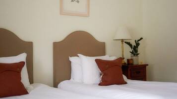 A hotel room with two beds, each with white bedding and brown pillows. There is a nightstand with a lamp and a small plant.