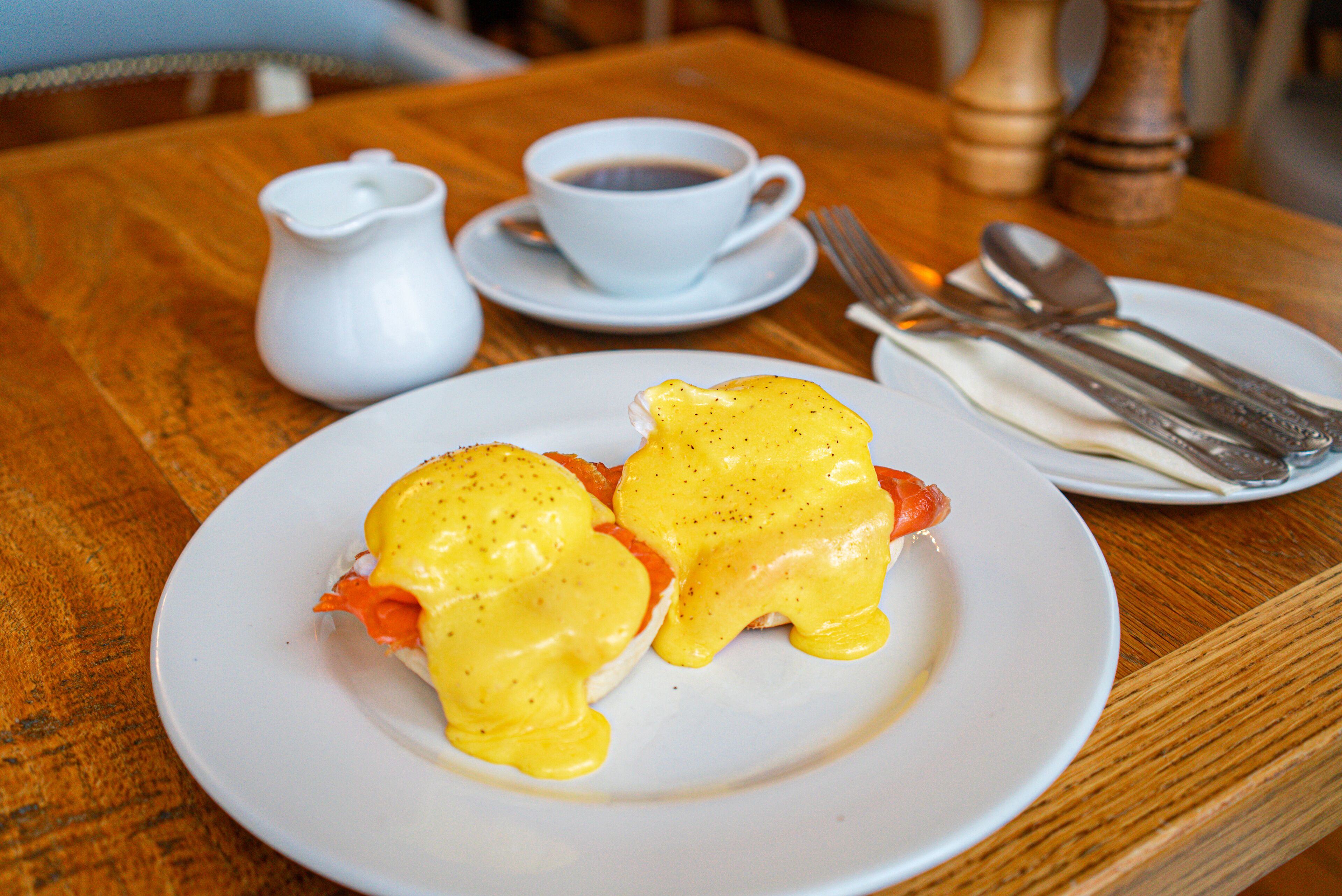 Uma mesa de madeira com ovos Benedict no prato, uma xícara de café e talheres.