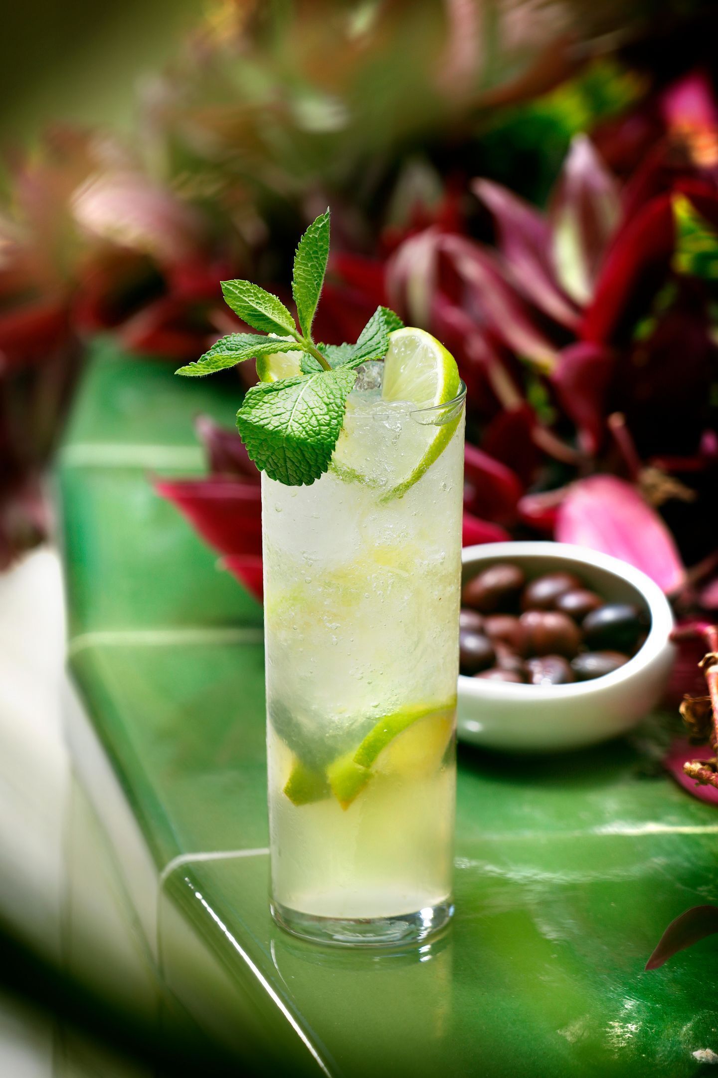 A tall glass of a lime and mint beverage on a green surface with a bowl of coffee beans in the background.
