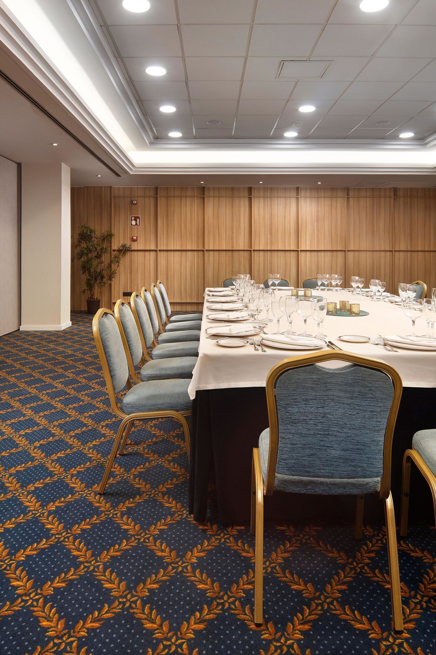 Uma sala de conferências com uma mesa comprida preparada para uma reunião, carpete estampado em azul e paredes revestidas de madeira.