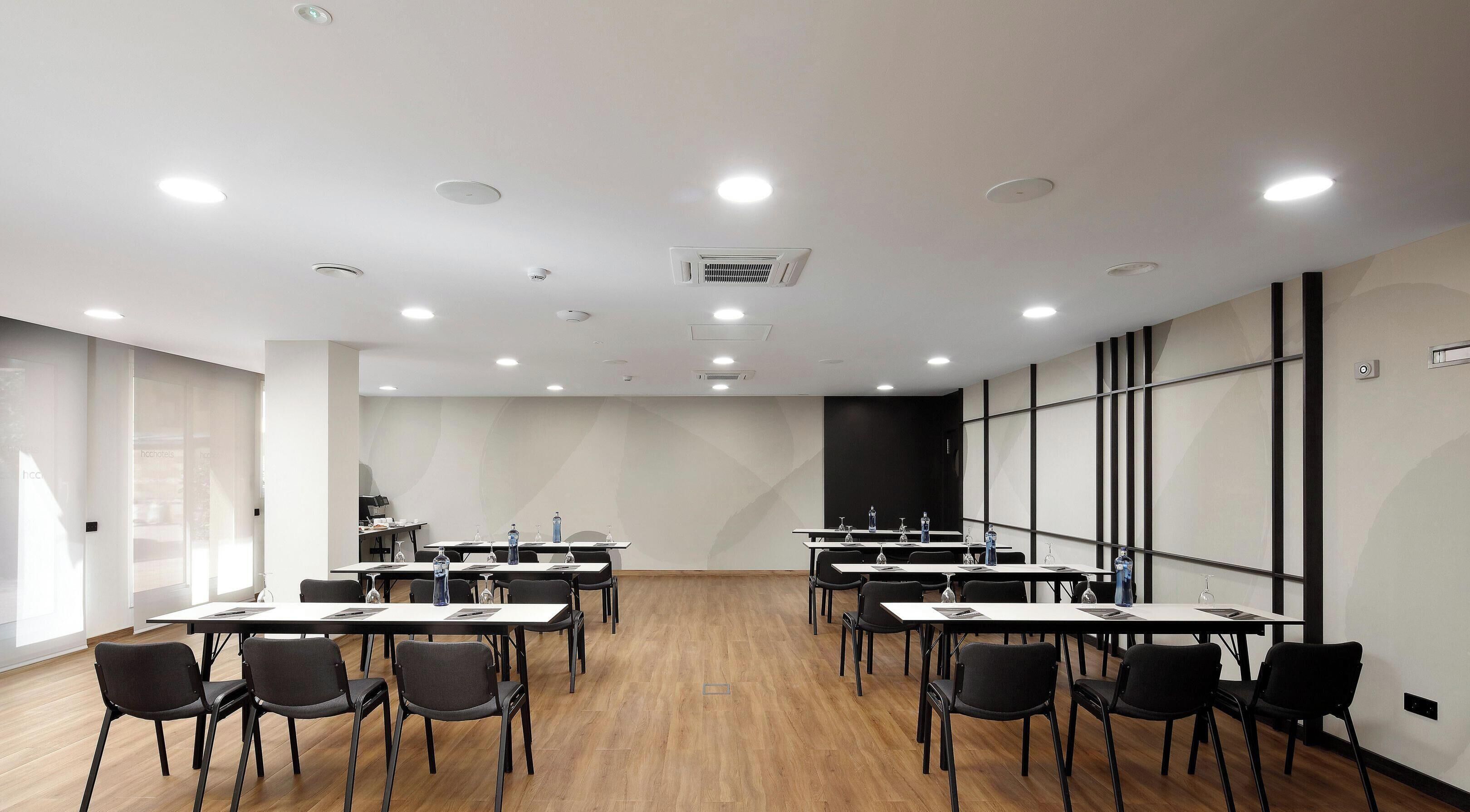 Sala de conferências com mesas e cadeiras, equipada com garrafas de água e uma grande janela lateral.