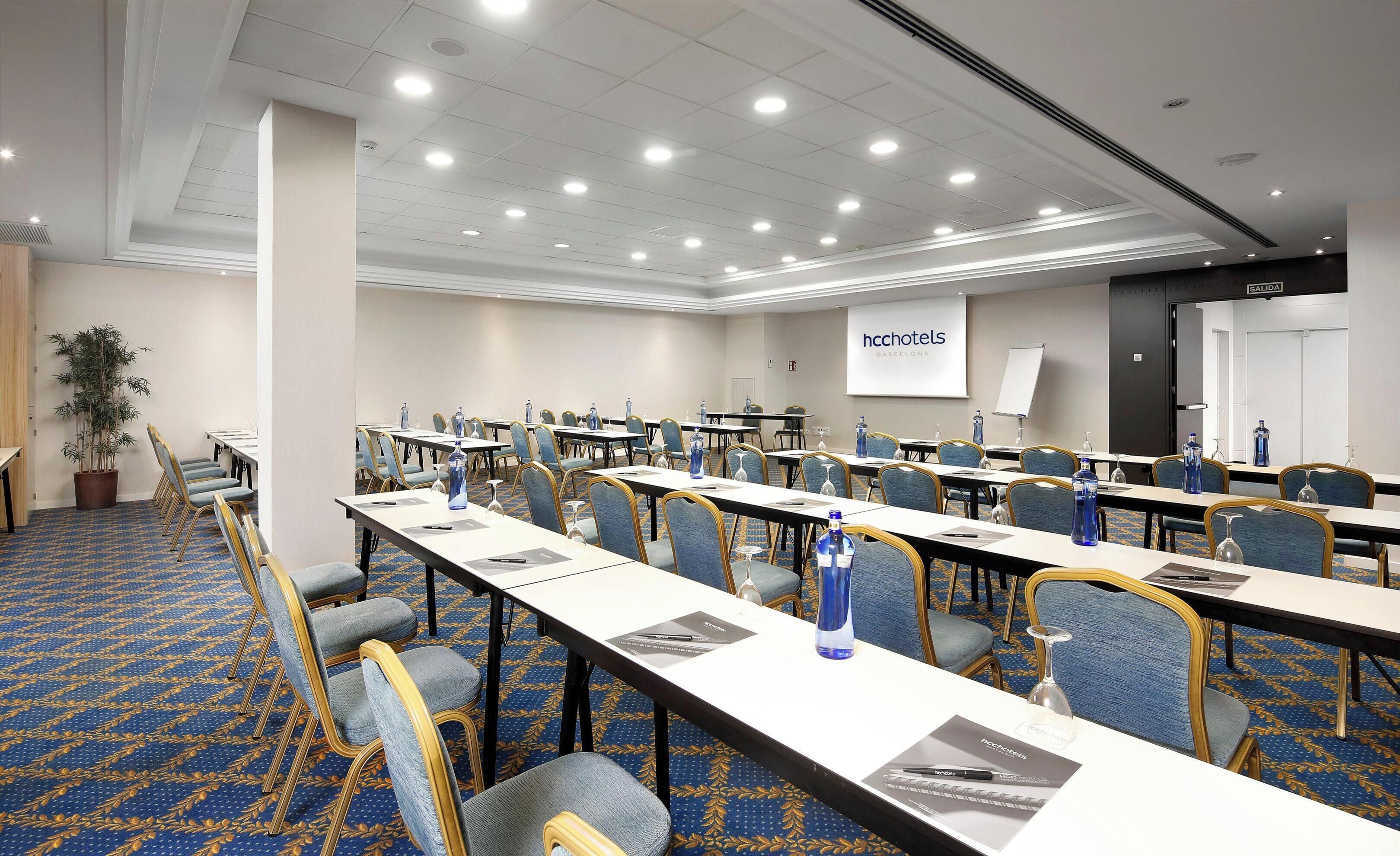 A conference room with rows of tables and chairs, a projector screen, and a sign for hcchotels Barcelona.