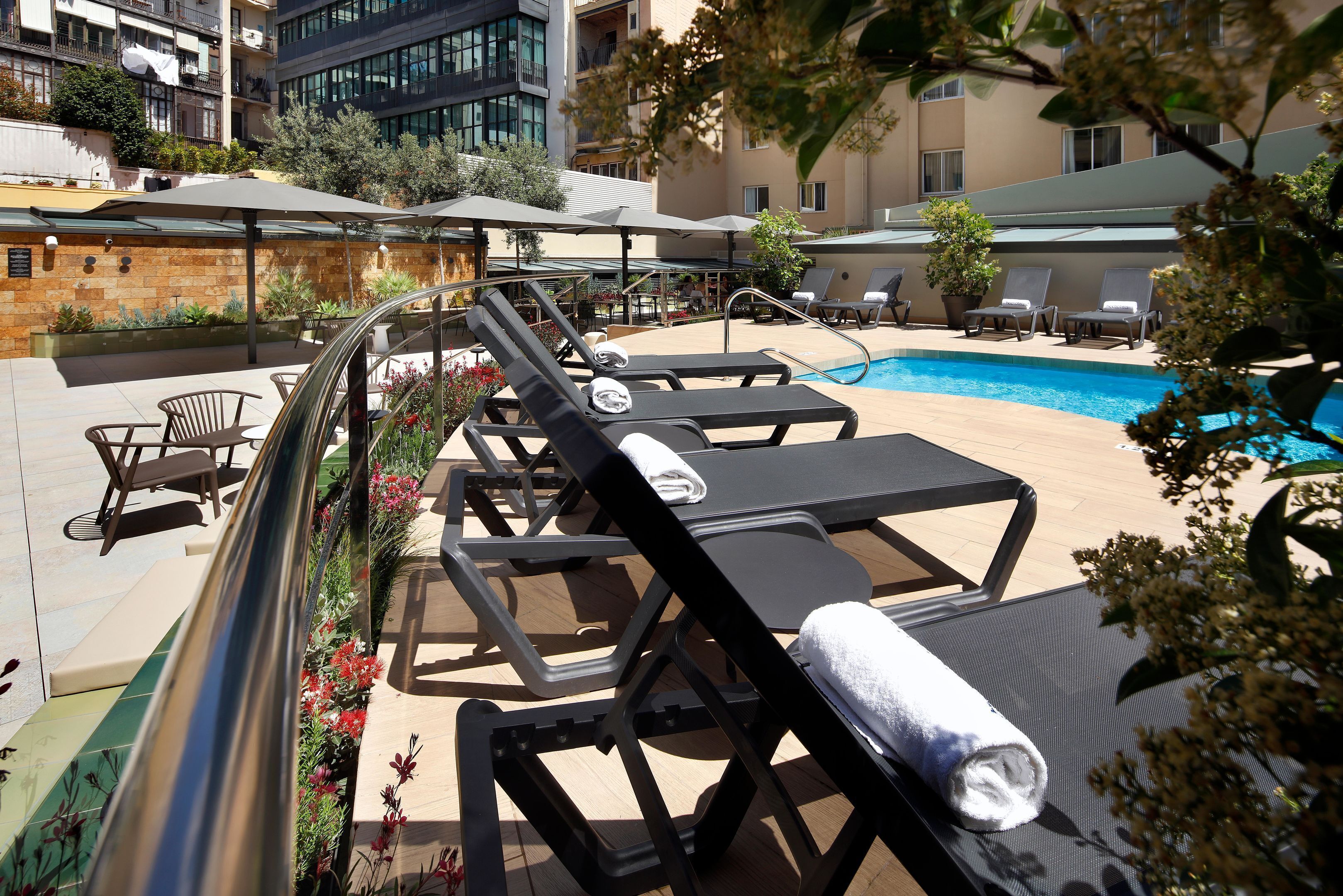 A rooftop pool area with lounge chairs and umbrellas, surrounded by a stone wall and greenery.