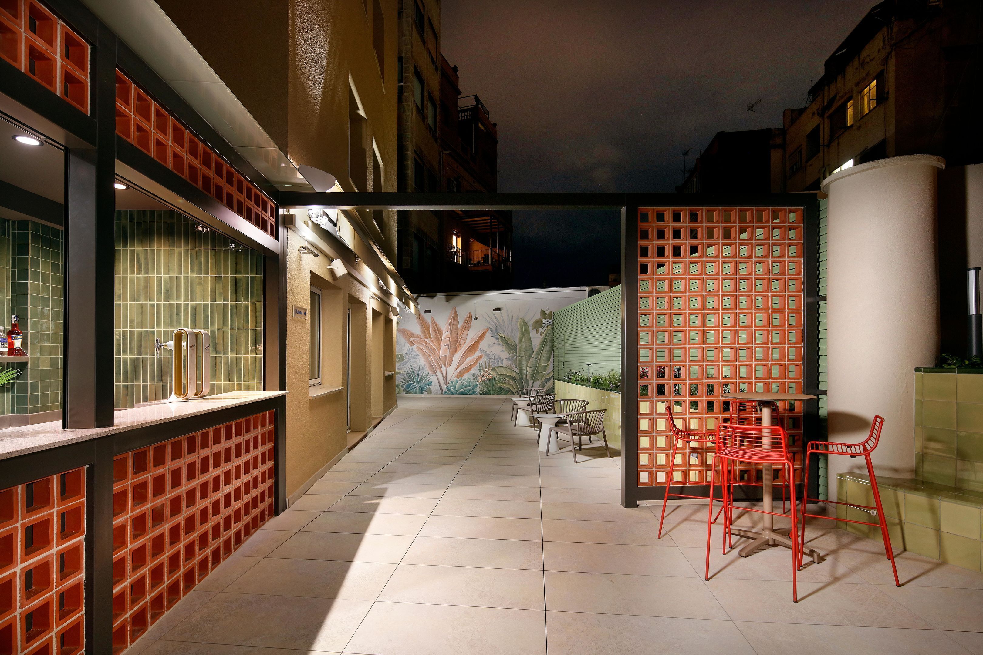 A narrow outdoor corridor with tiled flooring, a seating area with chairs, and a decorative wall with a leaf pattern.