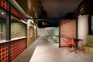 A narrow outdoor corridor with tiled flooring, a seating area with chairs, and a decorative wall with a leaf pattern.