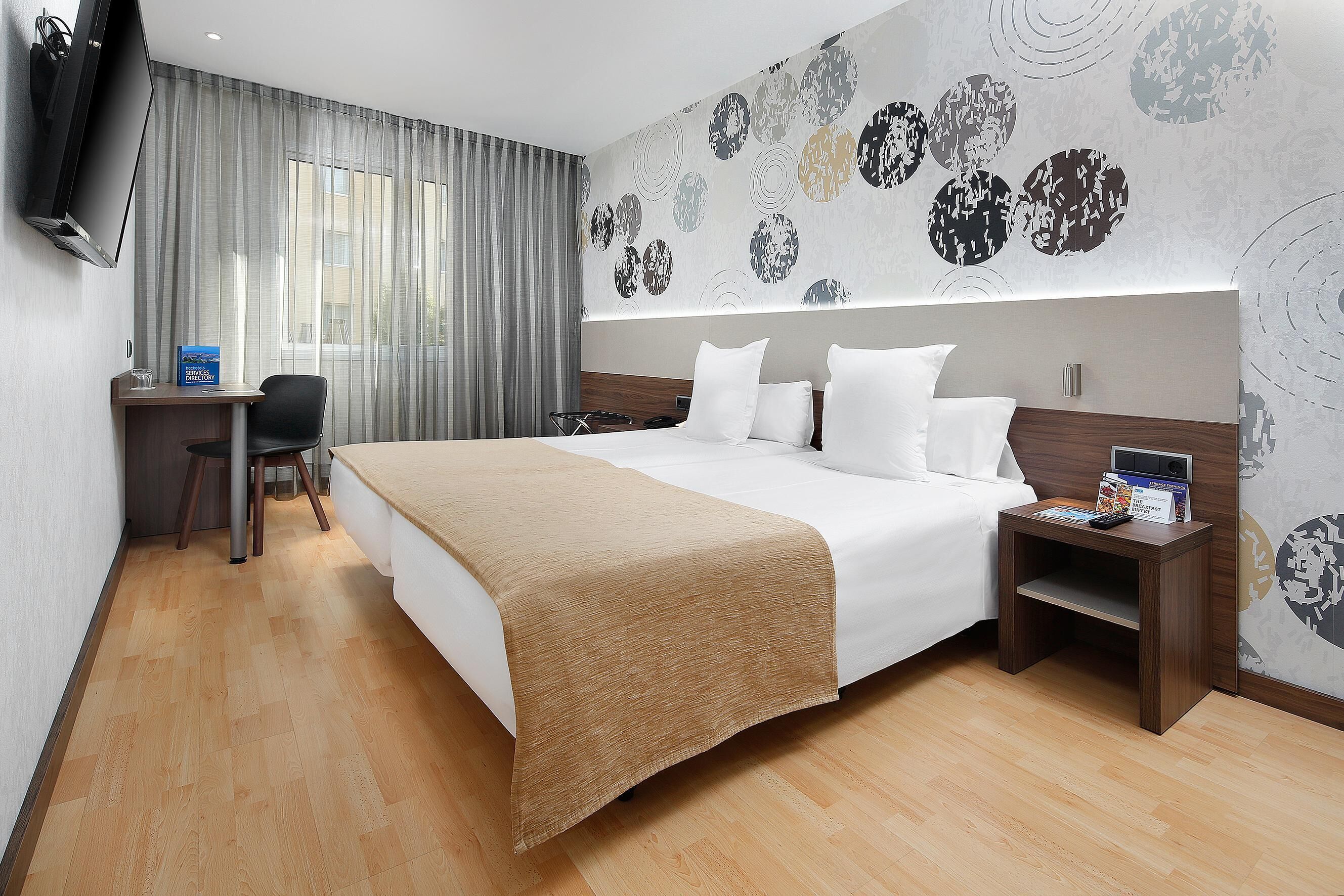 A hotel room with a bed, bedside table, TV, desk, chair, and a wall with a circular pattern.
