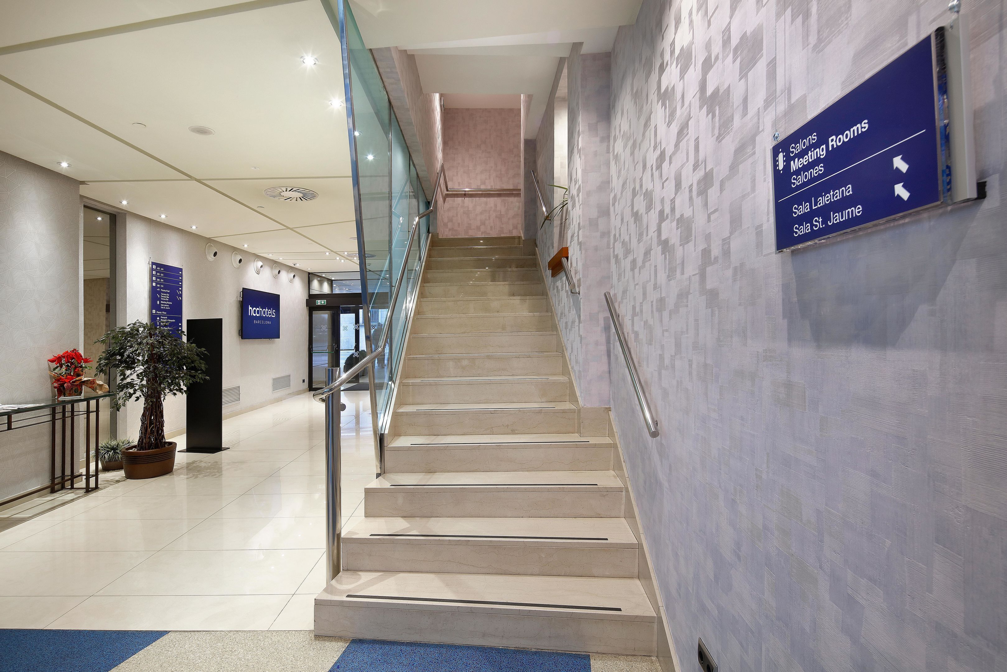 A modern hotel lobby with a staircase leading to meeting rooms, a reception desk, and directional signage.