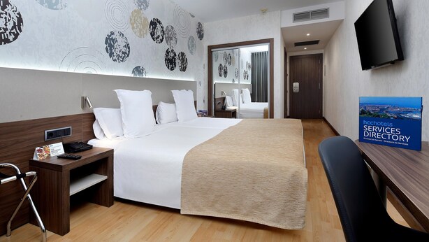 A hotel room with a large bed, a desk with a chair, a TV mounted on the wall, and a sign indicating hotel services directory. - Hotel HCC MontBlanc (Barcelona)